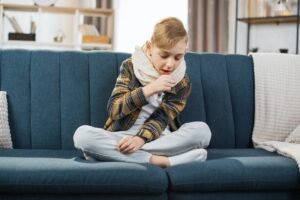 pleasant tired sick teen boy with scarf around neck sitting on soft couch and coughing